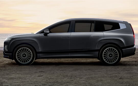A dark Hyundai Ioniq 9 shown in side profile, parked on a coastal overlook at sunset with large multi-spoke wheels and a streamlined SUV silhouette.