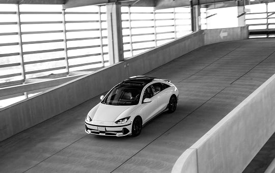 A white Hyundai Ioniq 6 driving through a multi-level parking garage, photographed from above in black and white.