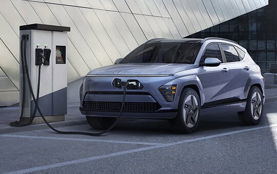 A light blue Hyundai electric SUV parked at a charging station, plugged in against a modern building backdrop.