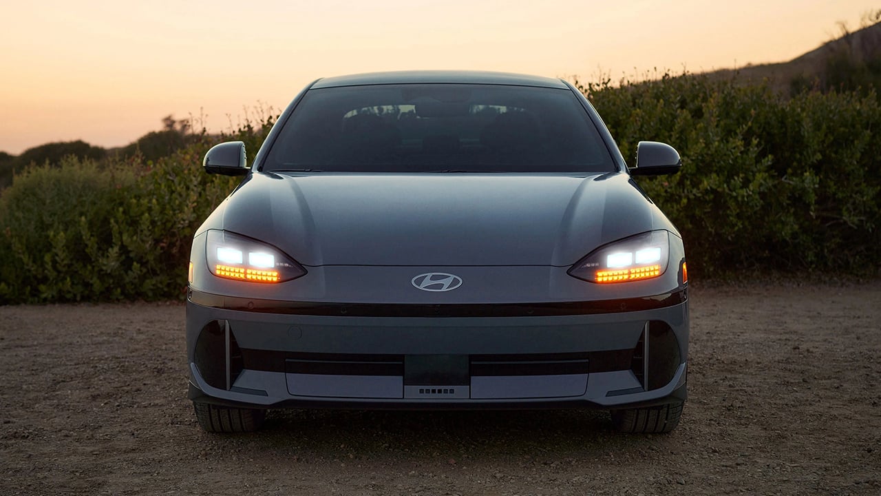 Vue avant du Hyundai Ioniq 6 2026 en arrêt sur route de sable avec phares allumées et couché de soleil