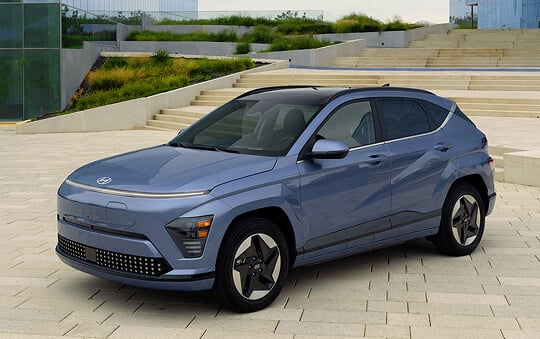 A light blue Hyundai Kona Electric parked on a paved plaza in front of wide concrete steps and landscaped greenery.