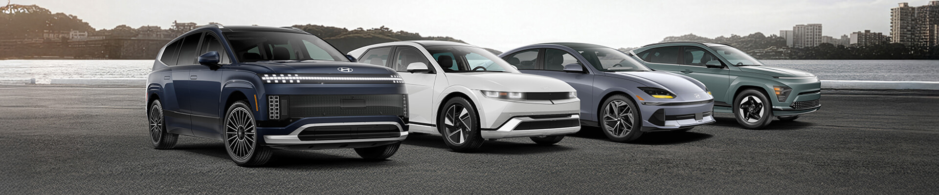 Four Hyundai electric vehicles parked side by side on a waterfront lot, with a dark blue SUV in front and three EVs in white, gray, and green behind it.