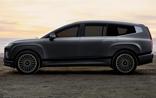 A dark Hyundai Ioniq 9 shown in side profile, parked on a coastal overlook at sunset with large multi-spoke wheels and a streamlined SUV silhouette.